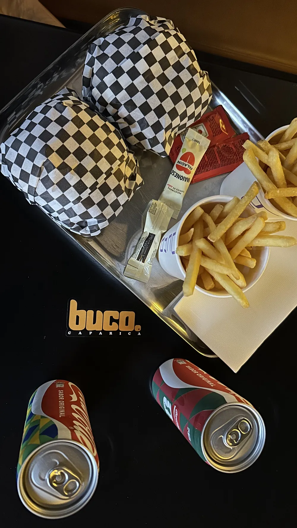 Two checkered burger boxes with fries and cans on a black tray